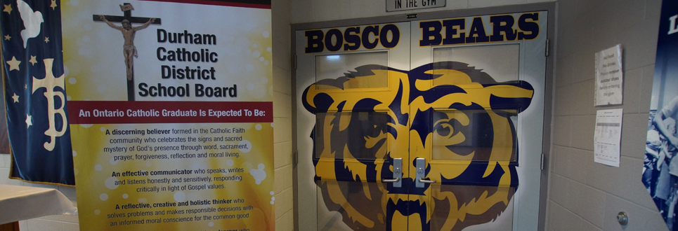 Stand-up sign of Catholic Graduate Expectations beside double doors painted with a giant grizzly bear face with BOSCO BEARS painted above it.